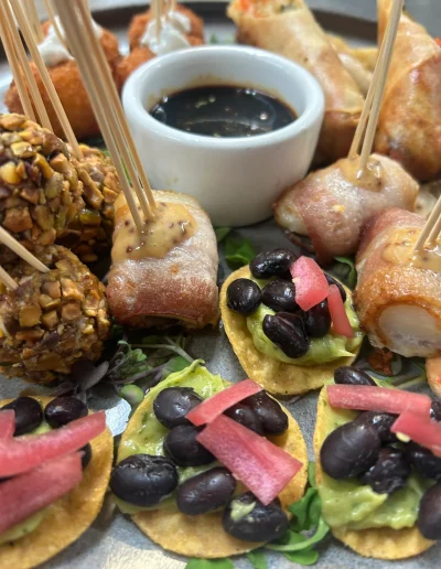 Assorted cocktail hour appetizers with bacon-wrapped bites, avocado black bean tostadas, pistachio-crusted bites, spring rolls, and dipping sauce