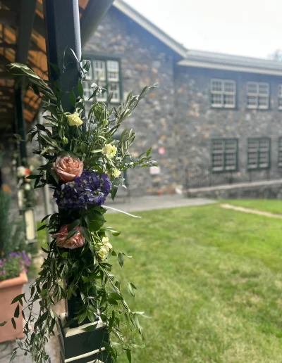 Floral column arrangement outside Rockwood Carriage House with pink roses, purple hydrangea, greenery, and stone building in the background