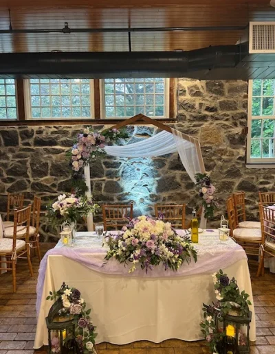 Sweetheart table setup at Rockwood Carriage House with floral arch, purple flowers, lanterns, and stone wall backdrop