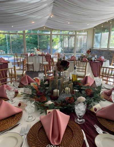 Wedding reception table setting at Rockwood Carriage House with pink napkins, burgundy runner, floral centerpiece, and draped ceiling